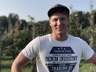 Junger Mann mit T-Shirt und Kappe in einem Garten mit Bäumen im Hintergrund.